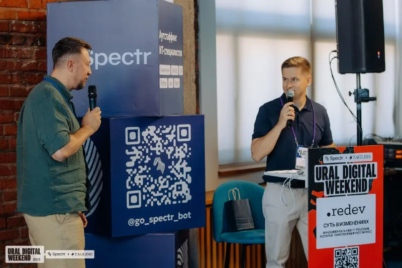 Александр Науменко выступает с микрофоном на сцене Ural Digital Weekend 2025 рядом со вторым спикером, на фоне стенды Spectr и Tagline